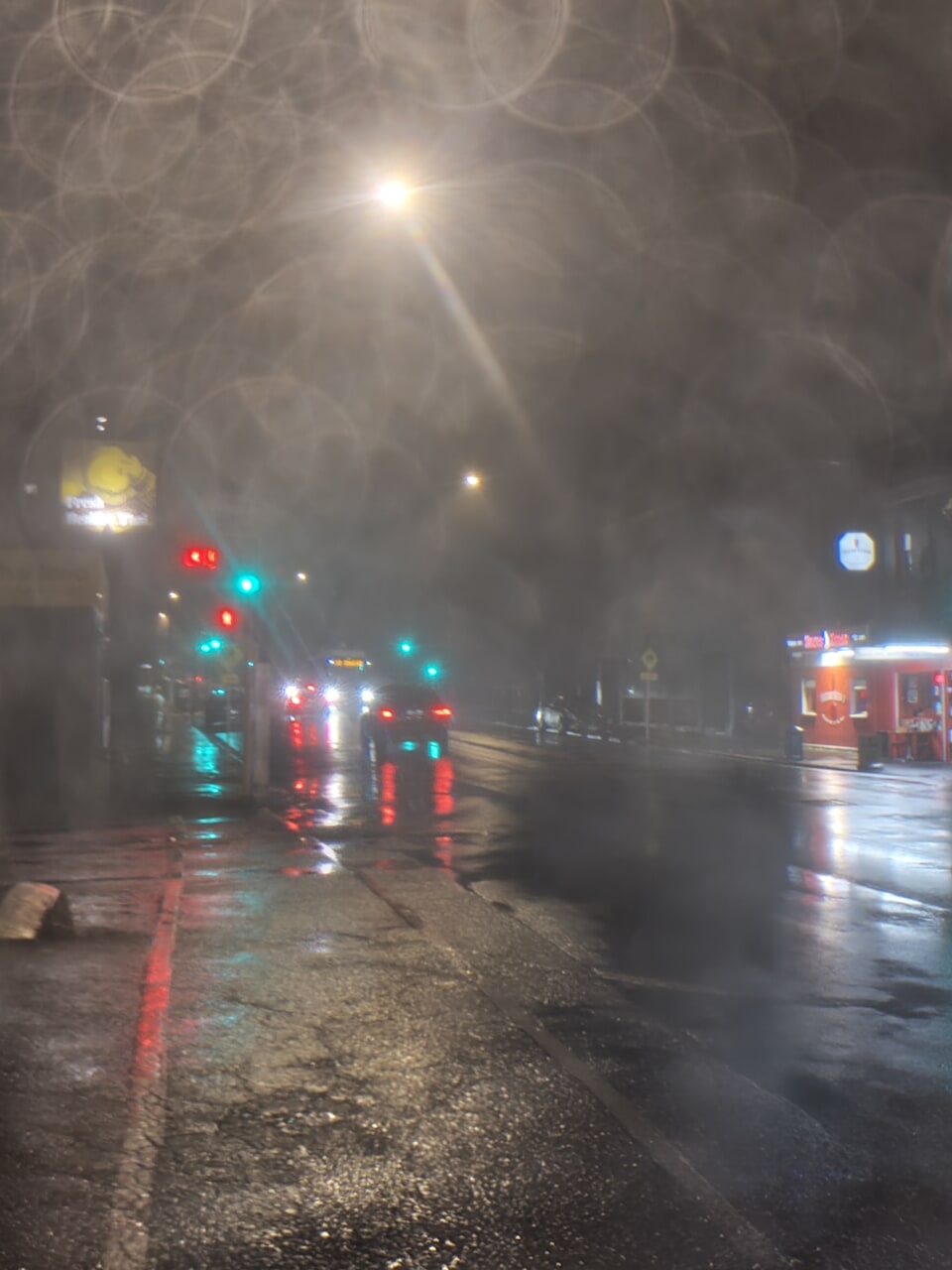 Extremely slimy and smudged looking photo of a rainy street at night. The bright lights form a stark contrast with the black air - or it would be black, if it weren't smudged grey by droplets.