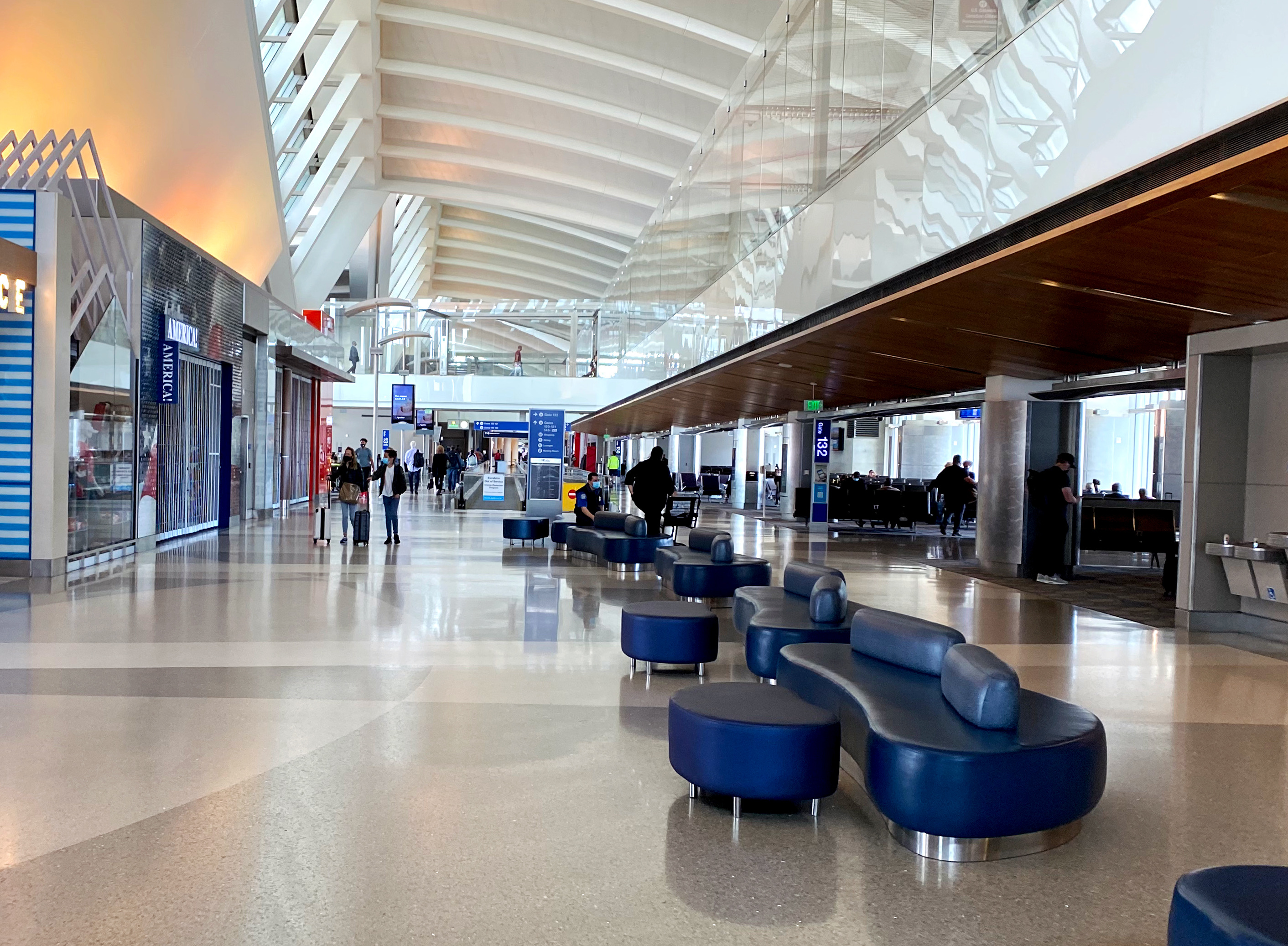 Looking down the main hallway of the terminal, with shopfronts on one side and gates on the other. In the middle of the hallway are some blue squishy benches, long enough to sleep on.